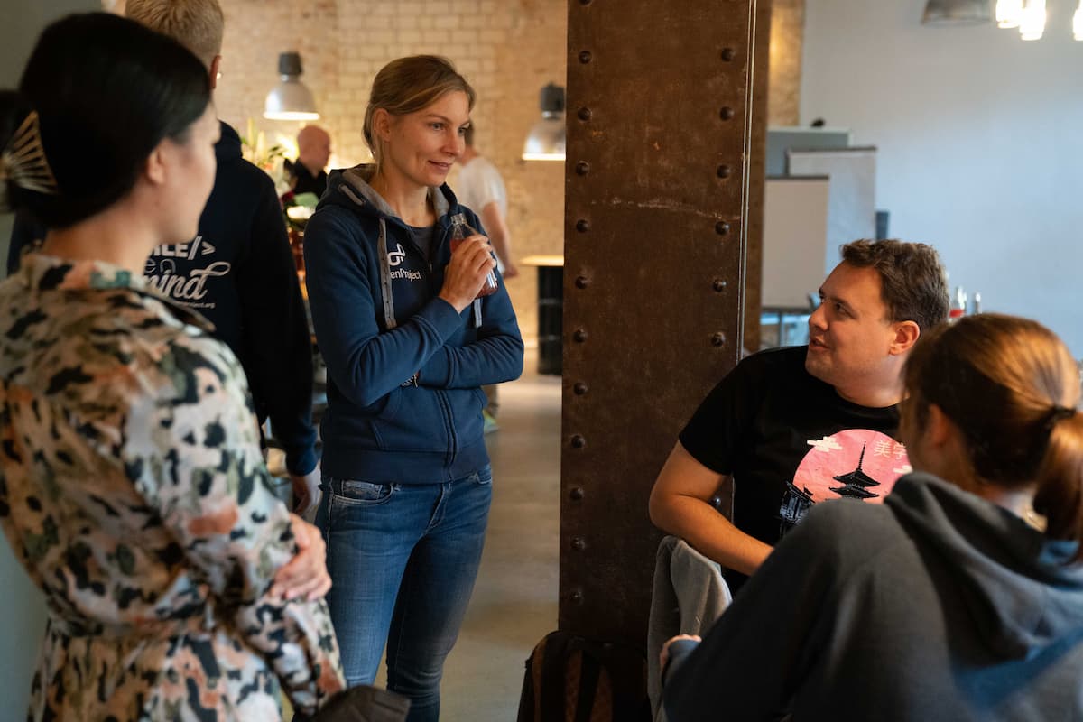 A man in a dark blue shirt sits at a wooden table, speaking with a woman. The background shows a blurred office space with other people and furniture.